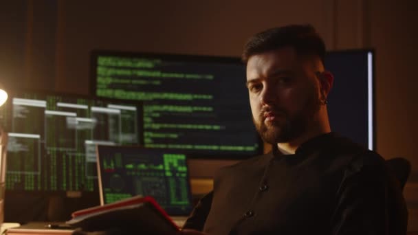 真剣な表情で若い黒髪の男のビュー 男性プログラマーは プログラムコードの実行行を持つモニターの背景にノートブックを手に彼の部屋に座っています — ストック動画