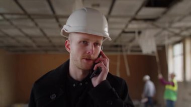 Bir bina müfettişi telefonda konuşuyor. Beyaz kasklı bir adam inşaat alanını kontrol ettikten sonra telefonla konuşuyor. İnşaat mühendisi birini arıyor. Yüksek kalite 4k görüntü