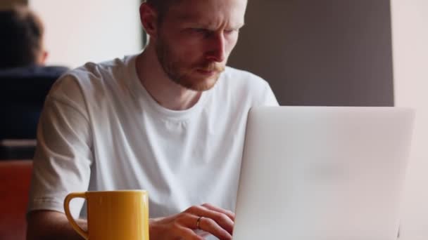 カフェに座っている間 真剣な男がノートパソコンのキーボードを入力しています 男はコーヒーを飲みながらカフェに座っている間 ノートパソコンでリモートで動作します 高品質4K映像 — ストック動画
