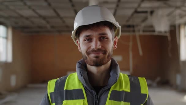 建設技術者はカメラに目を向け 微笑む ハードハットと反射ベストの陽気な建設労働者のフロントビュー 高品質4K映像 — ストック動画