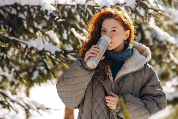 Gri ceketli kızıl saçlı bir kadın ısınmak için termostan çay içer. Kış kıyafetleri içinde genç bir kadın kış parkında yürüyor. Yüksek kalite fotoğraf