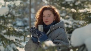 Karla kaplı ağaçların ortasında duran ve termostan çay içen güzel kızıl saçlı bir kadının yakın plan fotoğrafı. Sabahları karlı bir parkta yürüyen bir kadın termostan sıcak çay içer.