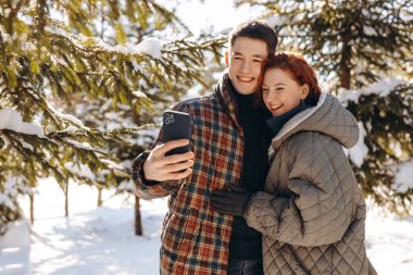 Mutlu genç bir çift, karla kaplı ağaçların önünde fotoğraf çekiyor. Gülümseyen gençler, karla kaplı bir parkta gündüz yürüyüşünde fotoğraf çekmeye karar verirler. Yüksek kalite fotoğraf