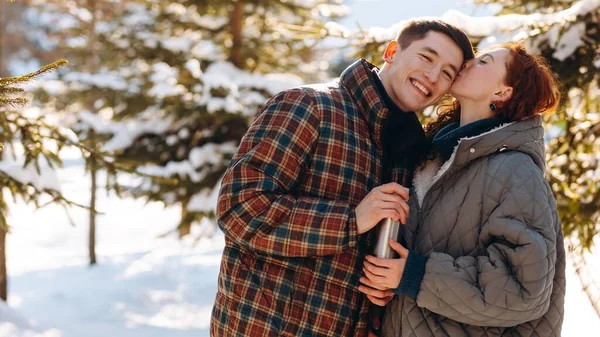两个年轻人站在雪地的森林中央 手里拿着热水瓶 一个美丽的红头发女人在冬天的森林里浪漫地散步时亲吻了她男朋友的额头 — 图库照片