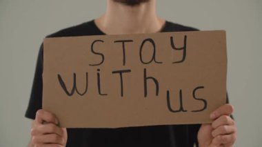 Adamın elinde tuttuğu görüntüler. Bizimle kalın. Gri bir duvara karşı duran erkek bir gösterici birlik ve destek çağrısında bulunuyor. Savaş karşıtı protesto. Yüksek kalite 4k görüntü