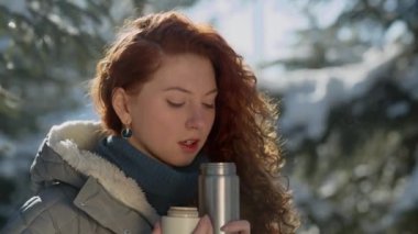 Kızıl saçlı bir kadının termostan sıcak çay içerkenki yakın çekimi. Kadın güneşli bir kış gününde yürürken sıcak çayla ısınmaya karar verdi. Yüksek kalite 4k görüntü