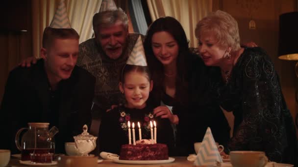 頭にお祝いの帽子を被ってテーブルに座っている女の子がケーキの上のろうそくを吹き消します 幸せな女の子はフレンドリーで愛情のある家族の輪の中で彼女の誕生日を祝う 高品質4K — ストック動画