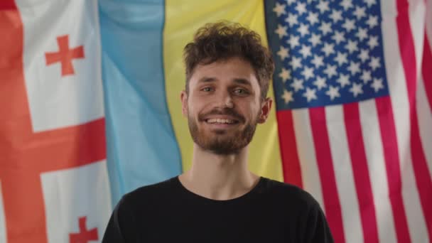 Ένας Νεαρός Χαμογελαστός Τύπος Στέκεται Στο Φόντο Των Σημαιών Της — Αρχείο Βίντεο