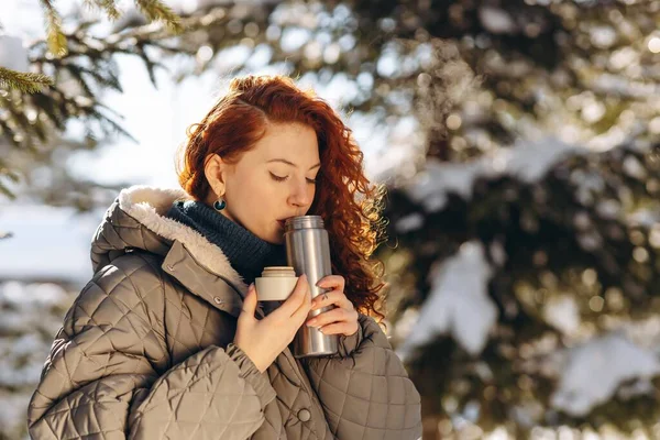 Termos bardağından su içen güzel bir genç kadın. Kızıl kızın kış günü parkta kendini sıcak çayla ısıtırken çekilmiş bir fotoğrafı. Yüksek kalite fotoğraf