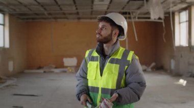 Kasklı inşaat işçisi elinde boya silindiri tutarak etrafa bakıyor. İnşaat alanında kasketli ve yansıtıcı yelekli bir mimar. Yüksek kalite 4k görüntü