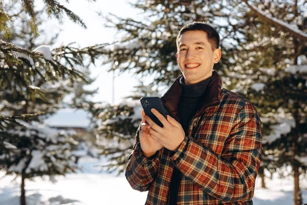 冬の森を背景に 携帯電話を手にした笑顔のブルネットの男がポーズをとっている 雪の真ん中に立っている再生ダウンジャケットの若い男のビューに覆われた木 高品質 — ストック写真