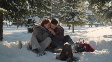 Kış ormanında oturan genç bir çift küçük bir ateşin yanında ısınmaya çalışıyor. Genç bir adam onları ısıtmaya çalışırken karısının kollarına nefes alıyor. Yüksek kalite 4k görüntü