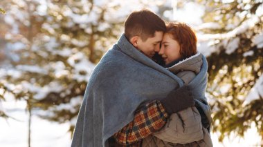 Güneşli bir ormanın ortasında battaniyeye sarılmış genç bir çiftin romantik bir fotoğrafı. Birbirine aşık bir çift, karla kaplı ağaçların arkasında birbirine tutunarak duruyor.