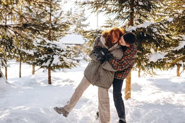 幸せな若いカップルは明るい晴れた冬の日に森の中を歩くのが楽しいです 若い男が自然の中を歩いている間 彼の赤い髪のガールフレンドを抱擁します 高品質の写真 — ストック写真