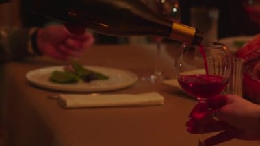 Bardağa kırmızı şarap döken bir adamın görüntüsü. İnsanlar kırmızı şarap içer ve bir restoranda akşam yemeğinde iyi vakit geçirir, bayramı kutlarlar. Yüksek kalite 4k görüntü