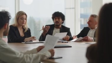 Takım elbiseli insanlar yönetim kurulu toplantısını konferans odasında yapıyorlar. Genç bir Hintli işadamı iş ortaklarına iş süreçlerini yeniden yapılandırma gereğini anlatıyor