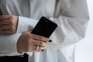 Beyaz gömlekli bir kadının elinde akıllı telefon tutuşu. Yüzük parmağında yüzük olan bir kadın elinde siyah bir akıllı telefon tutuyor. Yüksek kalite fotoğraf