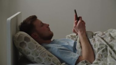 Genç bir adam yatakta uzanıyor ve akıllı telefondan sosyal ağlara takılıyor. Mavi tişörtlü bir adam yatmadan önce telefonundaki haberlere bakar. Yüksek kalite 4k görüntü