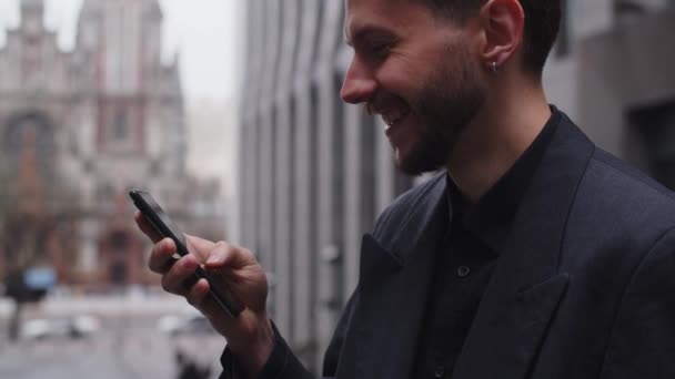도시의 배경에서 미소짓는 젊은이는 스마트폰을 정장을 수염을 거리에 전령을 누군가와 — 비디오