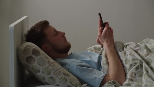 Młody Człowiek Leży Łóżku Przegląda Portale Społecznościowe Smartfonie Mężczyzna Niebieskiej — Wideo stockowe