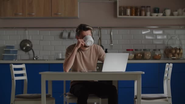 一个人在电脑前工作的时候 从杯子里喝着咖啡 一个坐在厨房里的男人正在笔记本电脑前努力工作 不时地从杯子里喝着咖啡 高质量的4K镜头 — 图库视频影像