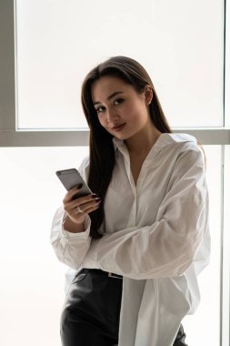Elinde telefon olan genç esmer bir kadın kameraya bakıyor. Beyaz gömlekli güzel bir esmer pencerenin arkasına poz veriyor. Yüksek kalite fotoğraf