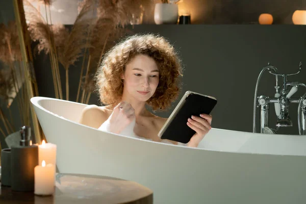 巻き毛の美しい女性が入浴中にデジタルタブレットを使用しています 泡風呂に座っている女性がデジタルタブレットでニュースフィードを閲覧しています 高品質の写真 — ストック写真
