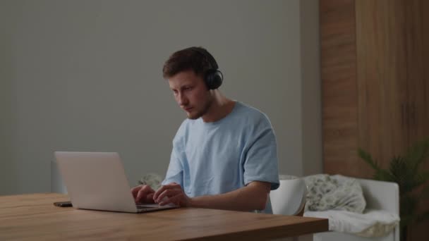 一个戴耳机的年轻人坐在笔记本电脑前的桌子前 在触摸板上滚动 一名年轻的男生坐在家里的电脑前搜寻网上的学习材料 高质量的 — 图库视频影像