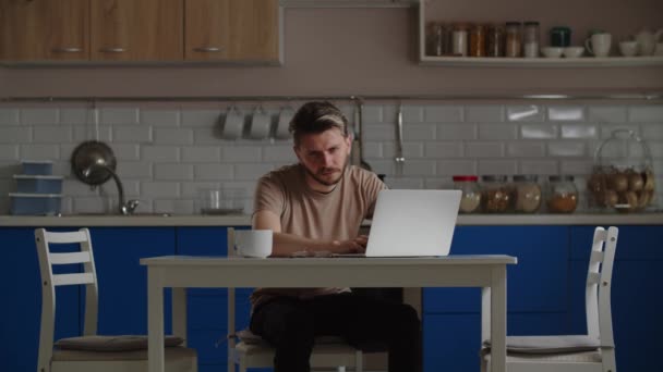 疲れた若い男が台所のテーブルに座り コンピュータ上の何かを見ている コーヒーを飲みながら座っている男性学生がノートパソコンでオンライン講義を聞く 高品質4K映像 — ストック動画
