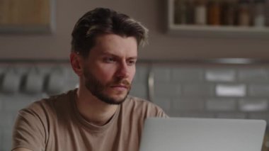 Bilgisayarda çalışmaya odaklanmış genç bir adam. Dizüstü bilgisayarın önünde oturan bir adam çenesini ovuşturuyor ve bir iş projesiyle sorunu nasıl çözeceğini çözmeye çalışıyor. Yüksek kalite 4k görüntü