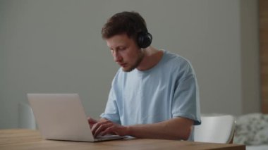 Dizüstü bilgisayar üzerinde çalışırken müzik dinleyen mavi tişörtlü bir gencin görüntüsü. Bir erkek öğrenci bir dizüstü bilgisayarda çalışma görevini yerine getirir ve kulaklıkla çevrimiçi bir konferans dinler. Yüksek kalite 4k