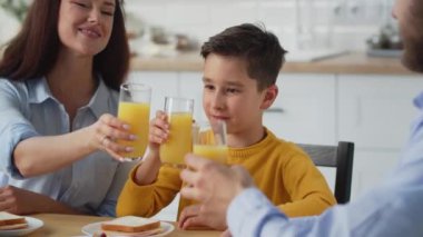 Oğlu portakal suyuyla bardakları tokuşturan gülümseyen aile. Çocuk ebeveynleriyle kahvaltıyı seviyor. Yüksek kalite 4k görüntü