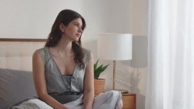 Genç, uzun saçlı bir kadın yatakta oturur ve pencereden dışarı bakar. Yeni uyanmış bir kadın pencerenin manzarasına hayran kalır ve bir şeyler düşünür. Yüksek kaliteli FullHD görüntüler