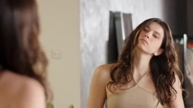 Tatlı bir esmer aynanın önünde duruyor ve kıvırcık saçlarını tarıyor. Bej kolsuz tişörtlü genç bir kadın saçını aynanın önünde tarıyor ve düzleştiriyor. Yüksek kaliteli FullHD görüntüler