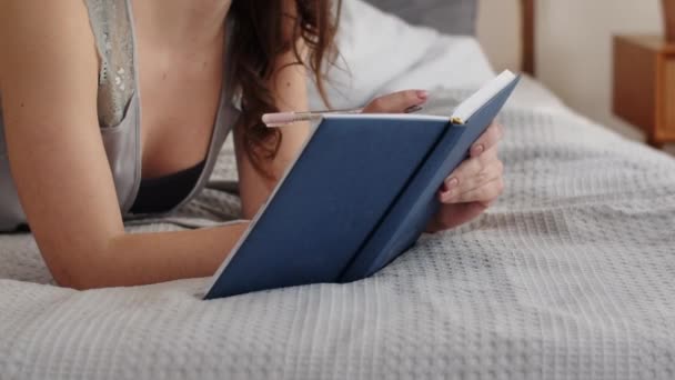 ベッドの上に横になってノートに青いカバーで何かを書いている女性の切り取られたビュー ブルネットは次の週のためのTo Doリストを作ります 高品質のフルHd映像 — ストック動画