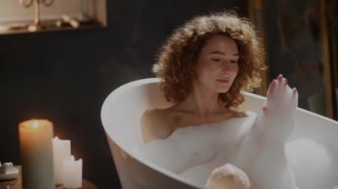 Kıvırcık kadın sıcak bir banyoda iyi vakit geçiriyor. Çekici gülümseyen kız banyo yapıyor ve yavaş çekimde kendine köpük sürüyor. Yüksek kaliteli FullHD görüntüler