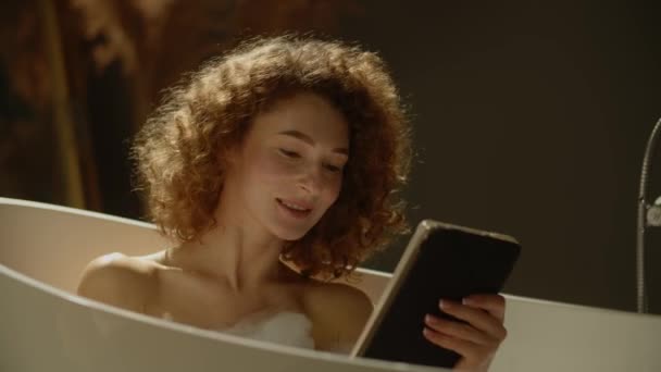 一位卷曲的女人正坐在泡泡浴中 通过数字平板电脑的视频链接与人交谈 一位女士洗澡时 通过视频链接与她的朋友讨论了上周发生的事情 — 图库视频影像