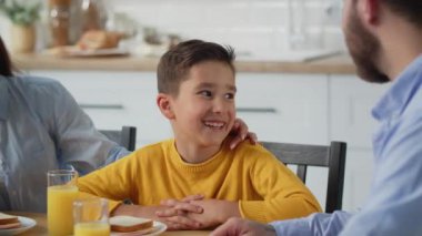 Gülümseyen bir çocuk anne ve babasıyla masada oturuyor. Çocuk kahvaltı sırasında mutfak masasında oturuyor ve ailesiyle bir şey tartışıyor. Yüksek kalite 4k görüntü