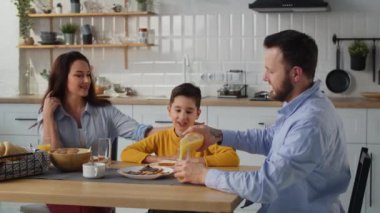 Sarı kazaklı bir çocuk mutfak masasında ailesiyle kahvaltı yapar. Masada oturan bir adam oğlu ve karısı için portakal suyu doldurur. Yüksek kalite 4k görüntü