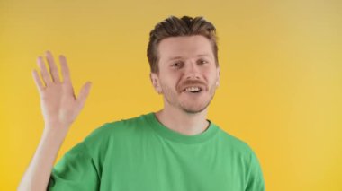 Yakışıklı genç adam el sallıyor ve sarı arka planda izole bir şekilde gülümsüyor. Mutlu adamın selamlaşması ön planda. Yüksek kalite 4k görüntü