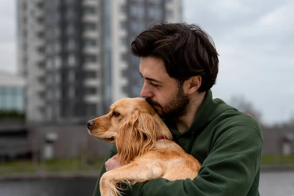 Brunette man holding his dog on blurred city background. Outdoor shot of a dog owner with his puppy. High quality photo