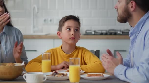 若い男は黙ってテーブルに座って 自分たちの間で何かを議論している彼の両親に耳を傾ける 黄色のセーターの少年が両親の間のテーブルに座ってカメラを見て — ストック動画
