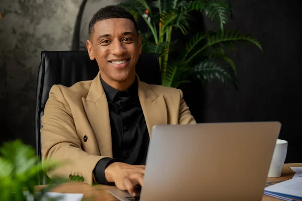 幸せなアフリカ系アメリカ人ビジネスマンの屋内撮影は 机の上にノートパソコンで作業 黒のビジネスオーナーのカメラに笑みを浮かべて 高品質の写真 — ストック写真