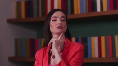 Kırmızı ceketli düşünceli esmer kadın kitaplığın yanında dururken bir şey düşünüyor. Yüksek kalite 4k görüntü