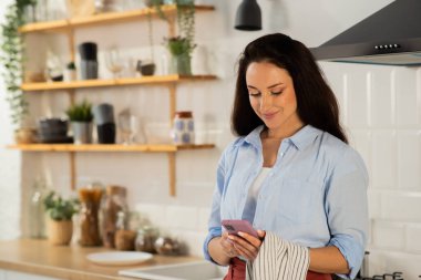 Güzel bir ev hanımı mutfakta duruyor ve akıllı telefon kullanıyor. Mavi gömlekli çekici bir esmer, kahvaltı hararetlenirken sosyal medya hesabına göz atıyor. Yüksek kalite fotoğraf
