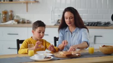 Güzel bir kadın mutfak masasında oturur ve oğlu için sandviç yapar. Sarı kazaklı küçük bir çocuk mutfakta annesiyle oturur ve sandviç yer. Yüksek kalite 4k görüntü