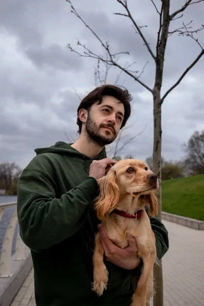 Genç bir adam bulutlu bir gökyüzüne karşı poz veriyor elinde bir cocker spaniel tutuyor. Yeşil ceketli esmer bir adam köpeğiyle birlikte kameraya poz veriyor. Yüksek kalite fotoğraf