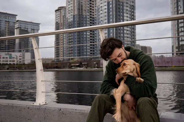 A young brunette man hugs a cocker spaniel and kisses him on the forehead. A man hugs his dog while sitting near the river against the backdrop of the city. High quality photo
