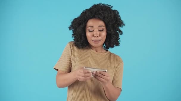 Μια Όμορφη Αφροαμερικανή Στέκεται Μπλε Φόντο Και Παίζει Στο Κινητό — Αρχείο Βίντεο