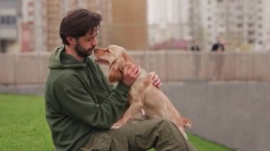 Bir adam parkta çimlerin üzerinde oturuyor ve kuyruğunu memnun bir şekilde sallayan bir cocker spaniel 'i okşuyor. Parkta bir gün yürüdükten sonra bir adam köpeğiyle çimenlerde oturur. Yüksek kalite 4k görüntü